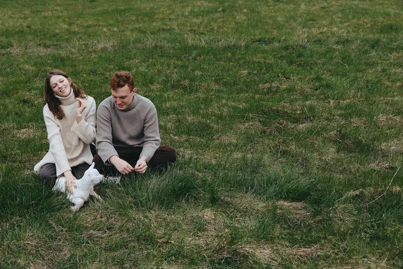 Young couple sits on grass in casual sweaters playing with a small white baby goat enjoying a relaxed lifestyle moment outdoors in early spring.