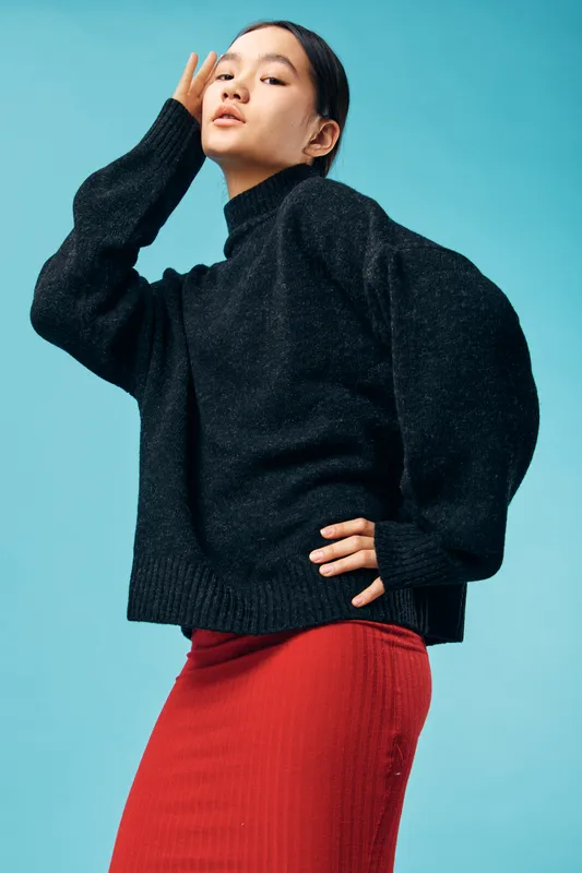 Woman posing in stylish black sweater and red skirt against blue background with confident expression and hand on hip for fashion and portrait photography concept.