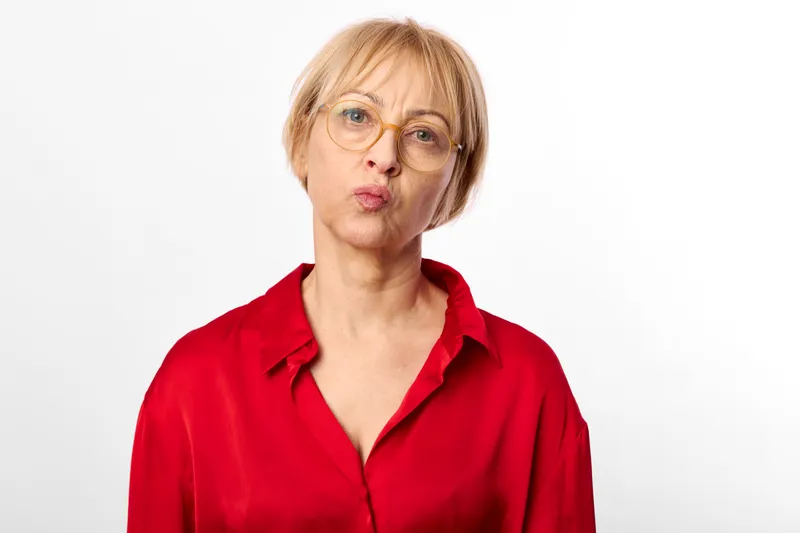 Senior woman with glasses and red shirt looks surprised expression, mouth slightly open, close portrait on white studio background, isolated mood, copy space, headshot