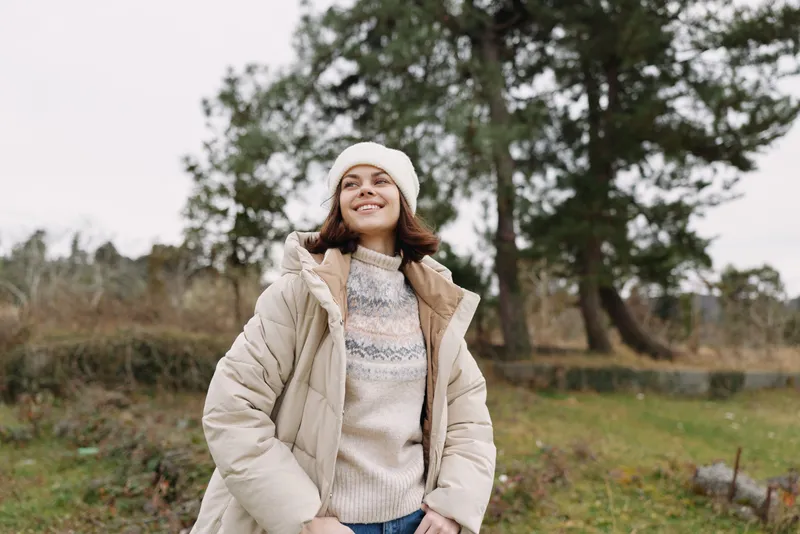 Young woman in a warm beige jacket and knit sweater stands outdoors with a beanie, smiling in a park setting during chilly weather and fresh air.