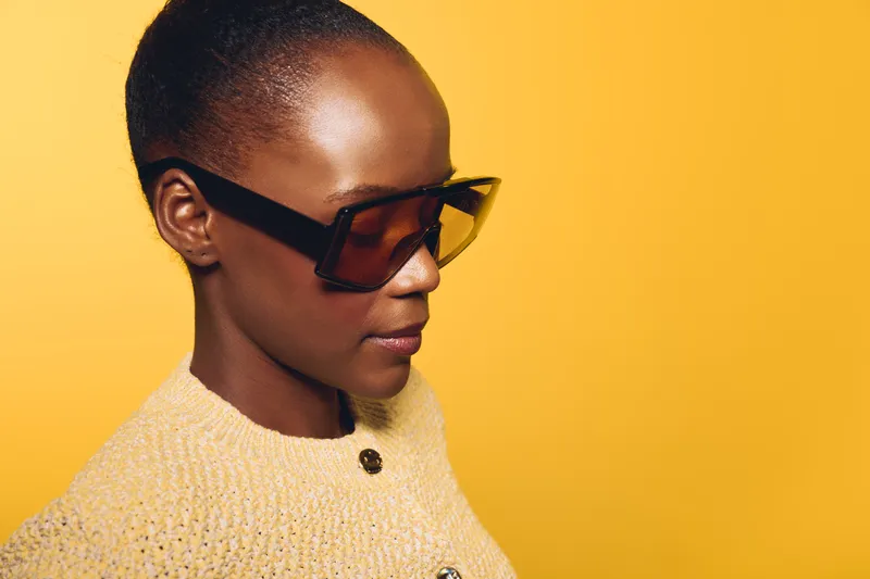 Portrait of a confident black woman with dark skin wearing large sunglasses and a beige knitted sweater against a bright yellow background. Concept of fashion and modern style.