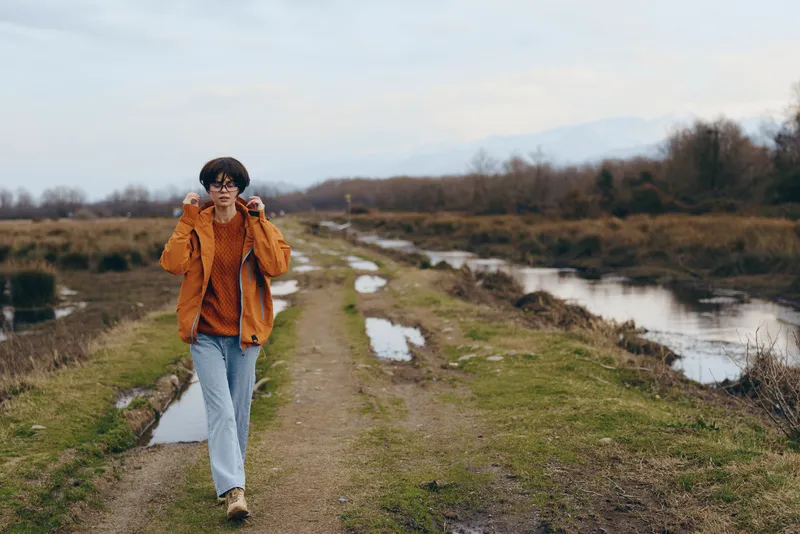 Woman lifestyle outdoors, hiking beside a river path in countryside. She wears an orange jacket and blue jeans, scarf and hat, walking confidently under overcast autumn sky quietly today