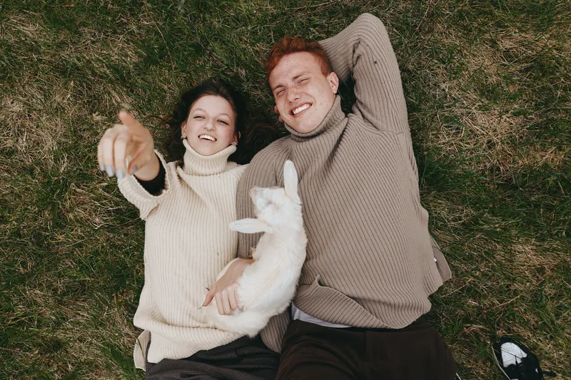 A young couple lies on the grass wearing cozy sweaters while holding a white baby goat in a relaxed outdoor lifestyle moment during autumn.