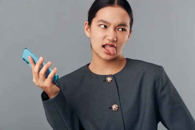 Woman in dark jacket holding smartphone with irritated expression looking to side, showing disgust with mouth open on gray background for communication and technology concept.