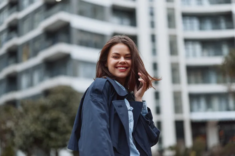 Smiling young woman with brunette hair wearing a denim jacket stands in front of modern city buildings, conveying confidence and urban style, with a bright, friendly mood.