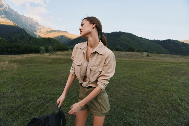 Young woman in a beige shirt and olive shorts stands in a wide meadow with distant mountains, turning her head as she adjusts a backpack for an outdoor excursion, conveying a candid moment of