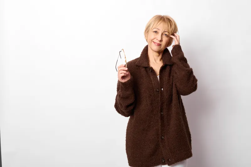 Woman blonde coat, studio portrait, smiling holding glasses near face, calm fashion mood, isolated on white background, minimal composition for text