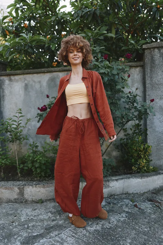 Young woman with curly hair wearing a rust jacket and matching pants twirls outdoors, radiating confident relaxed street style and a warm carefree mood. Soft natural light enhances her smile as green