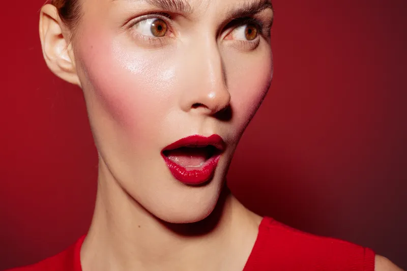 Close-up of surprised woman with red lipstick and blush on red background. Concept of beauty, makeup, fashion, and emotional expression in studio lighting.