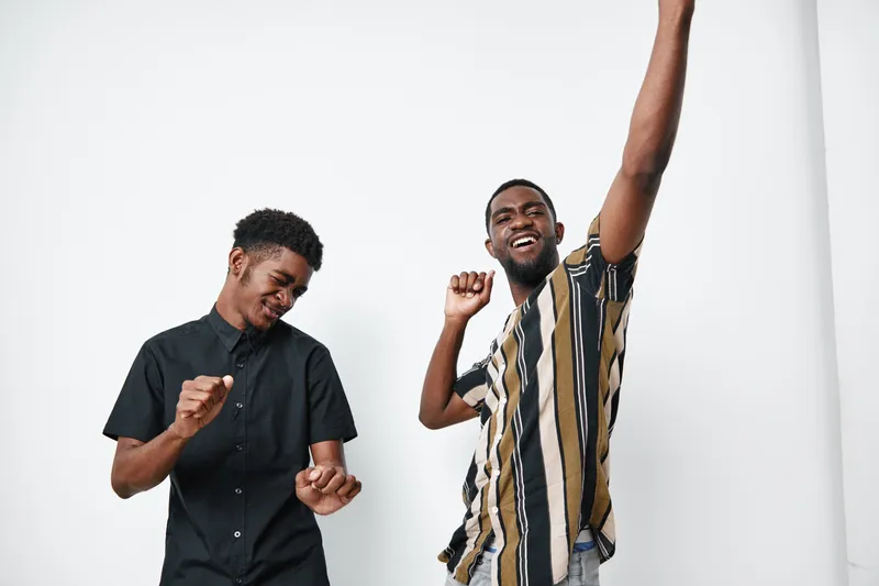 Two young black men with dark skin dancing and having fun together in casual clothes against white background. Happy friends enjoying music and celebration indoors.