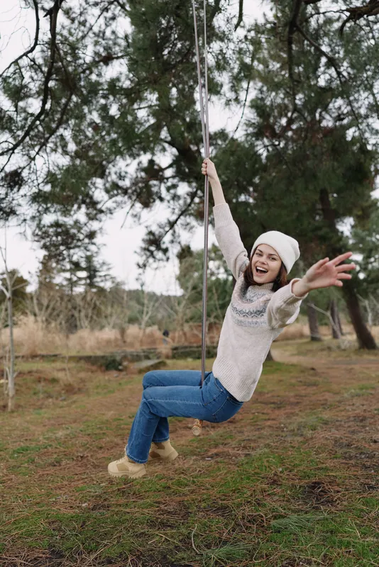 Young child on a swing in a park, outdoors, enjoying outdoor play, wearing a knit sweater and beanie, arms raised, smiling, carefree and full of fun, trees framing the scene.