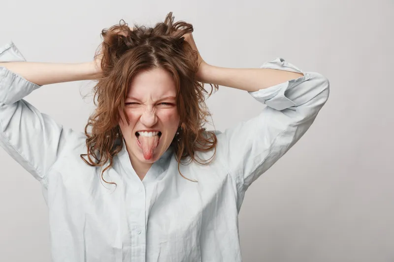 Young woman with curly hair wearing white shirt pulls her hair and sticks out tongue in playful expression. Studio portrait on light background. Concept of fun, emotions, and youth.