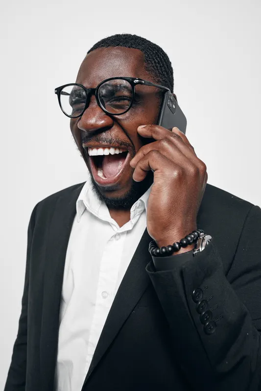 Black man with dark skin wearing glasses and suit talking on smartphone with excited expression isolated on white background. Communication, technology and emotion.