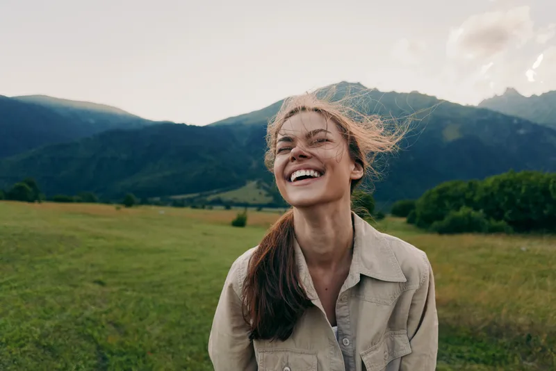 A young woman with long hair smiles in a sunlit field with distant mountains, conveying carefree outdoor happiness and natural vitality.