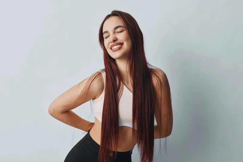 Young woman with long braided hair wearing sportswear smiling with closed eyes and hands on lower back in studio. Healthy lifestyle, fitness, wellness and relaxation concept.
