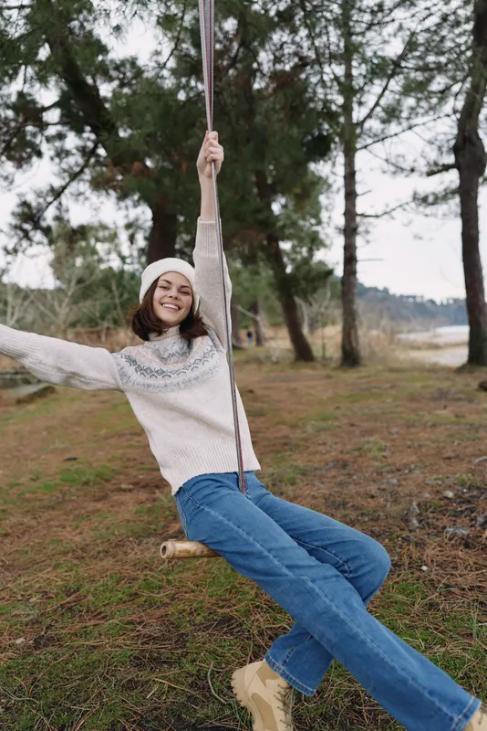 Outdoor swing scene with a smiling girl enjoying a casual moment in a park, trees in the background, cozy sweater, jeans, and wooden seat captured in natural daylight