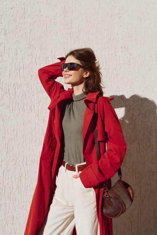 Young woman in red coat and sunglasses smiles confidently standing against textured wall with hand in hair enjoying sunny day outdoors.