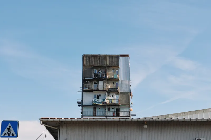 Apartment building with stacked balconies and weathered facade under a blue sky, urban housing scene showing character and decay along a quiet street