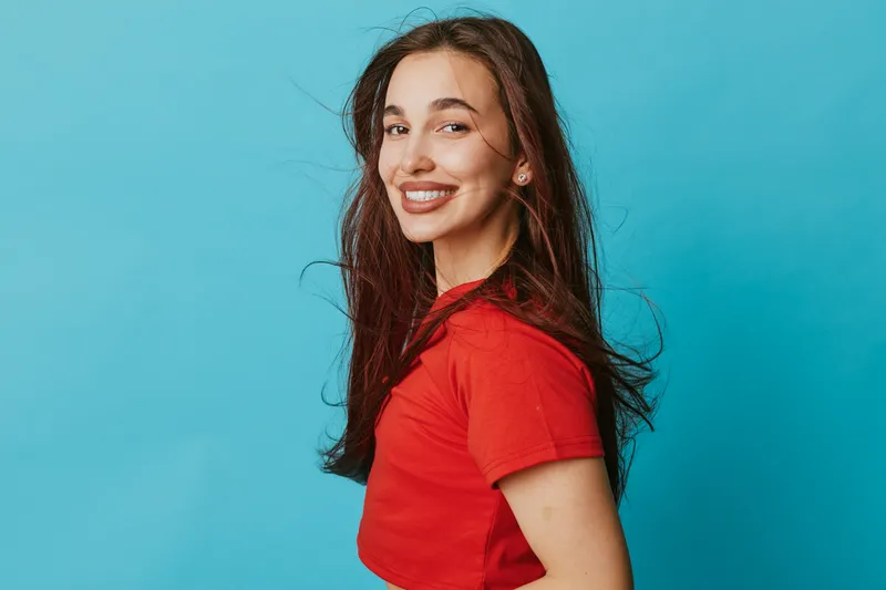 Young woman smiling and posing in red t shirt with long hair against blue background for portrait and positive emotion concept in studio lighting.