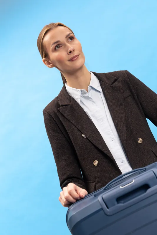 Young businesswoman in formal jacket stands with suitcase on blue background looking thoughtful. Concept of travel planning, business trip, and professional lifestyle.