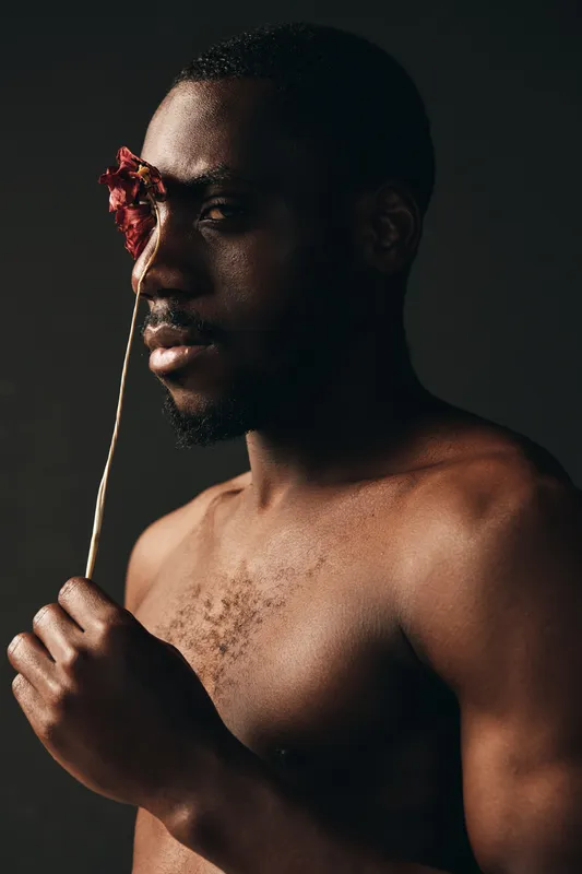 Creative conceptual portrait of African man with dark skin holding dried flower near face in studio. Artistic visual style with bare chest and moody lighting for emotional expression.