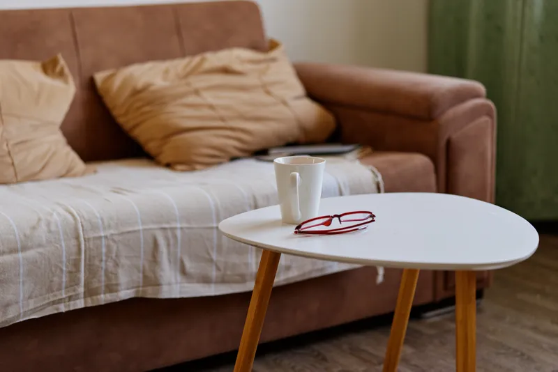 Cozy living room scene with a light sofa, small round table, mug resting nearby, soft natural light, relaxed home lifestyle, warm neutral tones, minimal decor