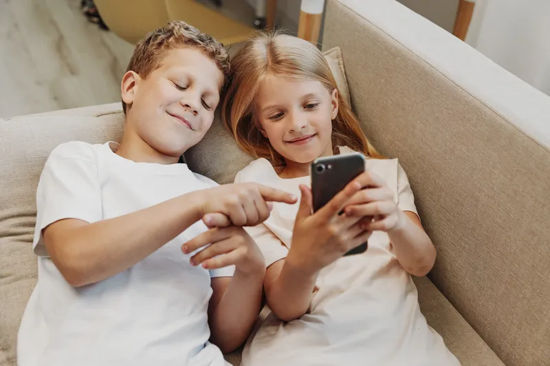 Two smiling children sitting on a couch looking at a smartphone together, sharing moments of joy and curiosity in a cozy home setting.