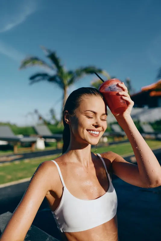 A smiling woman in a white bikini top enjoys a cold red drink by the pool under clear blue sky. Palm trees and lounge chairs create a relaxed summer lifestyle atmosphere.