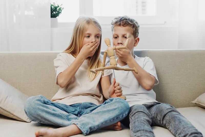 Two children sitting on a sofa holding a wooden mannequin, both covering their mouths in playful surprise. Bright natural light fills the cozy living room setting.