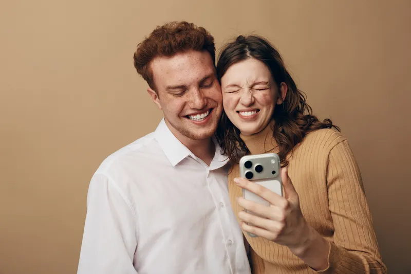 Happy young couple takes a selfie together smiling and laughing. Casual clothing and warm beige background create a cozy and joyful atmosphere. Concept of friendship and love.
