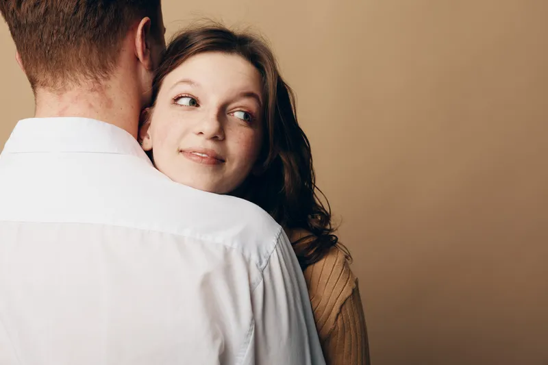 Young woman with brown hair embraces man wearing white shirt, looking sideways with gentle smile. Intimate moment of affection and connection in warm neutral background setting.