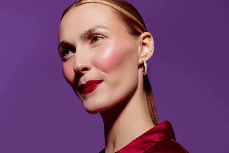 Close-up portrait of confident woman with shiny skin and red lipstick against purple background. Concept of beauty, skincare, makeup, and fashion style in studio lighting.