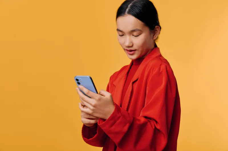 Young woman in red jacket holding smartphone and texting message, standing against yellow background. Asian female using mobile phone for communication and social media interaction.