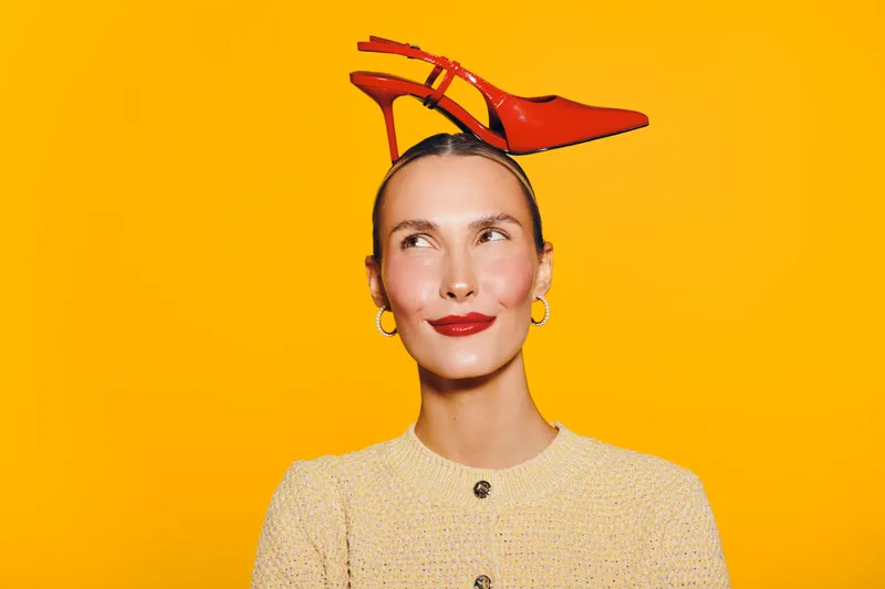 Young woman with red high heel shoe on her head looks up smiling against bright yellow background. Concept for fashion, creativity, and playful style with vibrant colors.