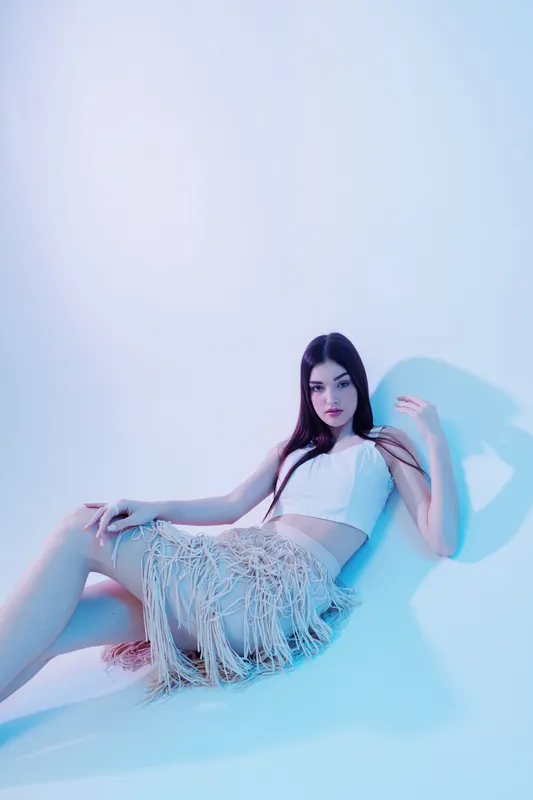 Young woman in a white crop top and fringed skirt poses seated on a light background. The image highlights beauty, fashion, and modern style concepts.