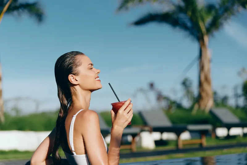 Relaxed lifestyle of a woman by a pool enjoying a refreshing drink, sunny outdoor resort setting, wet hair, white swimsuit, serene pose, leisure mood, sparkling water, tropical ambiance.