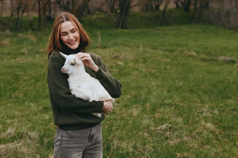 Cheerful woman outdoors, holding a goat kid in a green field, wearing a cozy sweater, soft smile, gentle breeze, natural colors, pastoral landscape, lifestyle moment, warm and inviting