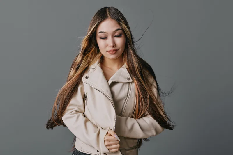 Young woman with long hair wearing light jacket posing with arms crossed against gray background for fashion style and portrait photography, beauty and casual look.