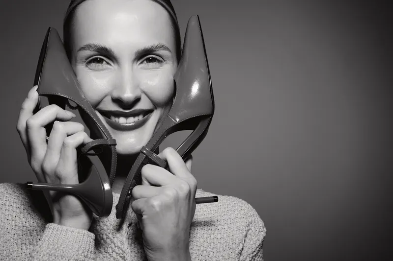 Smiling Caucasian woman with light skin holding high heel shoes near face in studio, expressing joy and fashion concept.