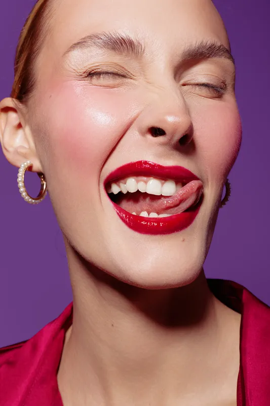 Close-up of a smiling woman with red lipstick and hoop earrings winking and licking lips. Bright purple background and vibrant red blouse create a playful and confident mood.