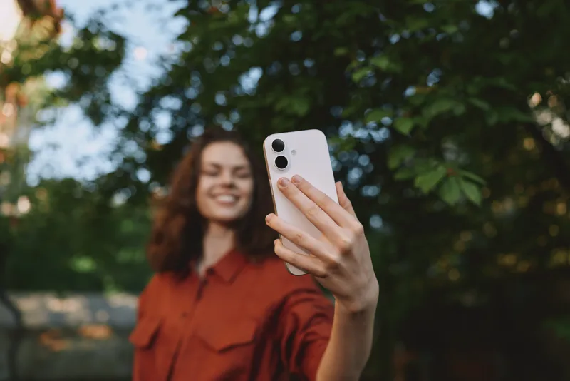Young woman with curly hair in a rust shirt takes a selfie outdoors, smiling at her smartphone against a leafy background