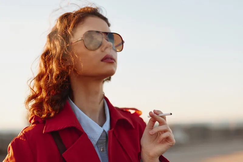 Confident woman in a red coat and sunglasses outdoors, smoking a cigarette, stylish street fashion portrait with a soft sunset backdrop and calm urban mood today.