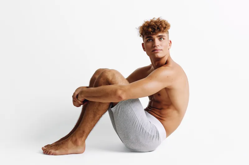 Young man with curly hair sitting on floor in shorts against white background, posing shirtless with relaxed expression and crossed arms around knees in studio.