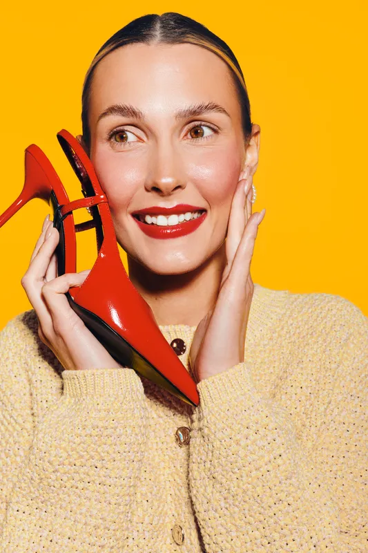 Smiling woman with red lipstick holds red high heel shoe near face on bright yellow background. Concept of fashion, style, beauty, and joyful emotions in studio lighting.