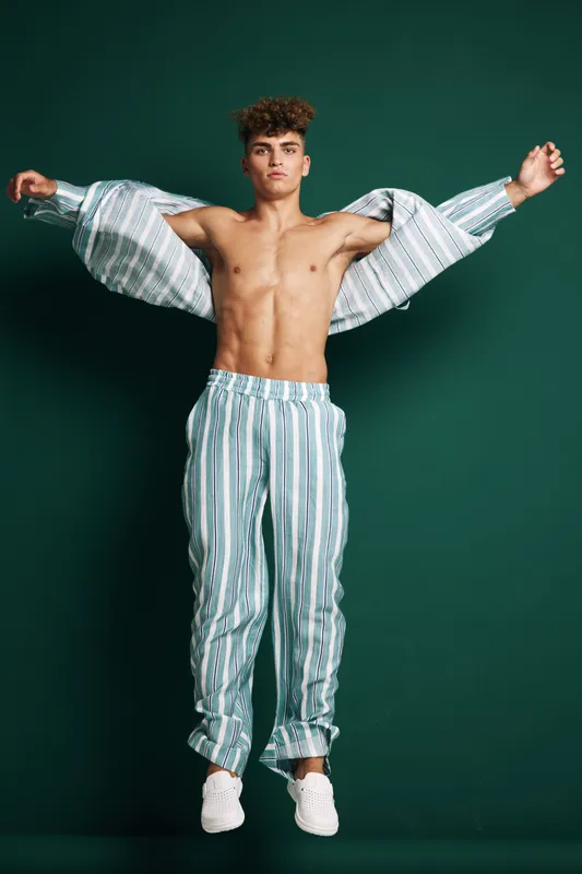 Young man with curly hair wearing striped pajama pants and open shirt standing against dark background with arms raised and bare chest visible in studio lighting.