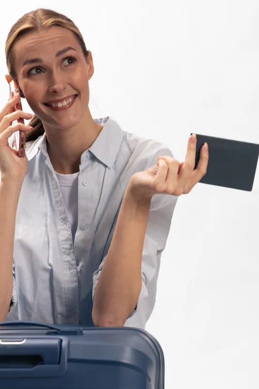 Smiling young woman holding passport and credit card while talking on phone near suitcase. Concept for travel planning, booking, and vacation preparation with happy tourist in studio.