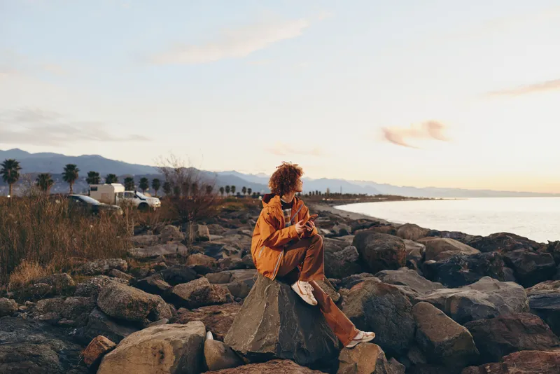 Woman lifestyle on rocky coast at sunset, warm golden light on curly hair, ocean waves in distance, traveler posing outdoors, peaceful calm nature mood, seaside travel