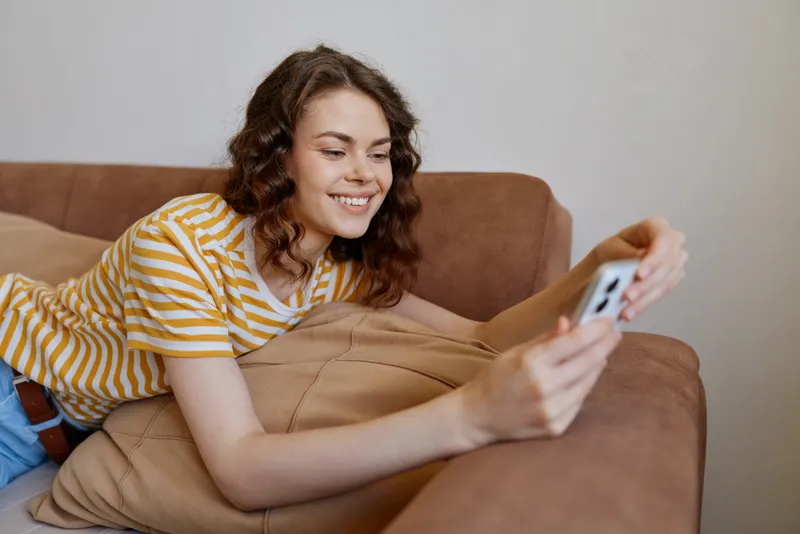 Cheerful woman relaxing at home on a couch, smiling while filming a selfie with a phone, striped tee, casual pose, warm natural light, cozy living room atmosphere, candid lifestyle moment