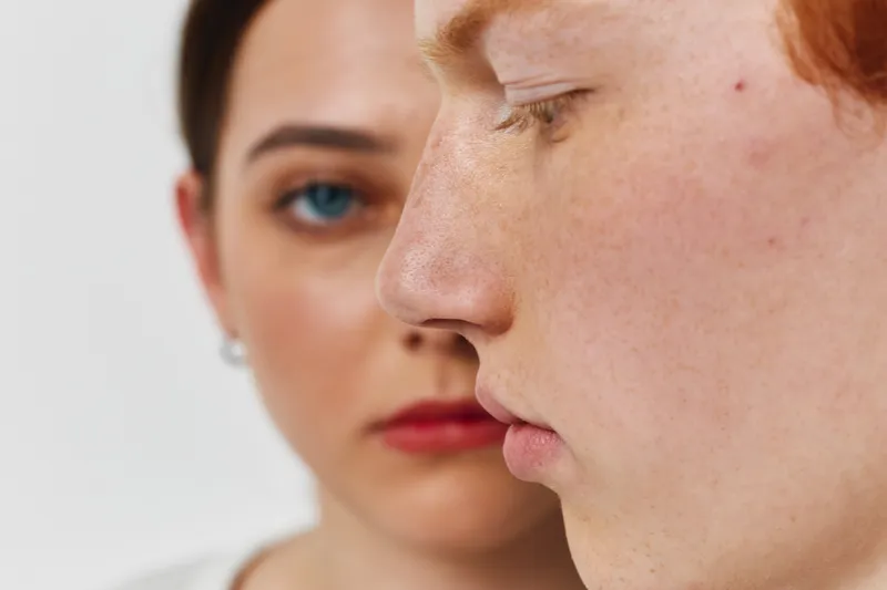 Two young people with closeup faces showing skin texture and expression, one focused on profile with freckles and the other blurred in background with blue eyes and red lips.