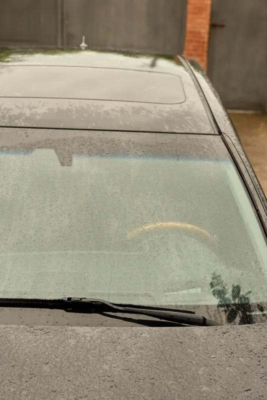 Close-up of a rain speckled windshield with a resting wiper on a wet hood, urban driveway setting, moody reflections, everyday travel atmosphere